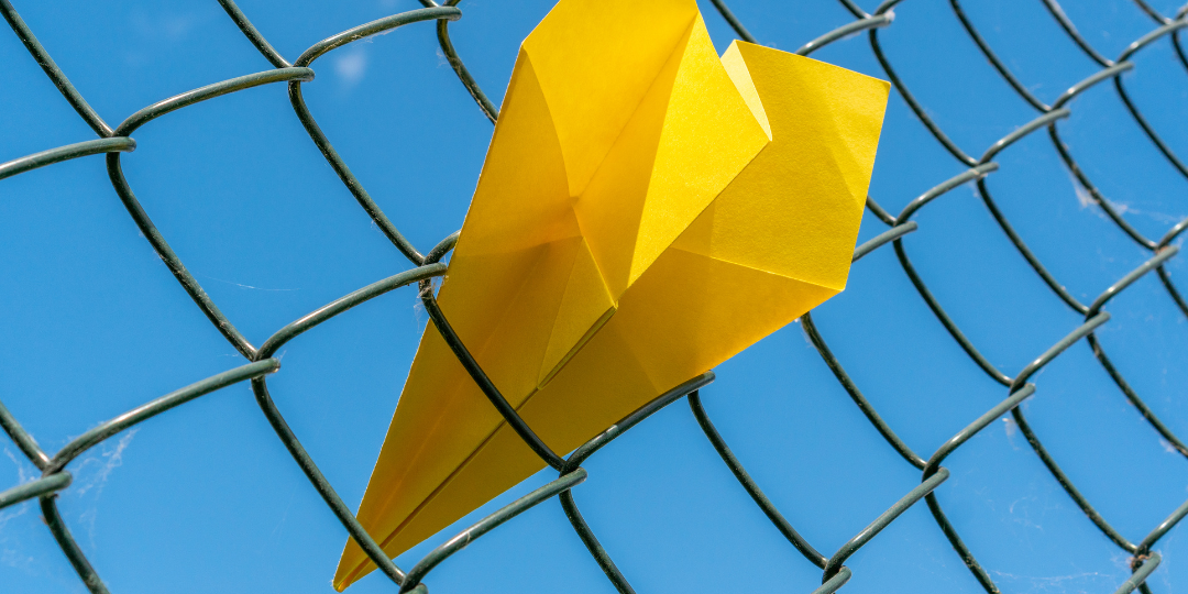 Yellow paper airplane stuck in a chain-link fence against a blue sky, representing being stuck in anxiety