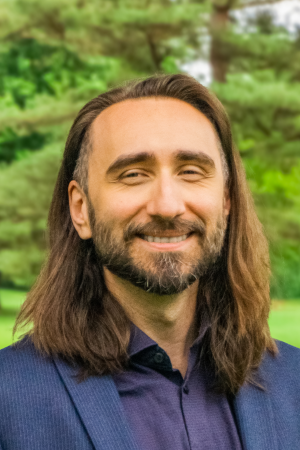 Headshot of Dr. Alexey Breuss, a man with long brown hair and a beard, smiling warmly against a blurred green background.