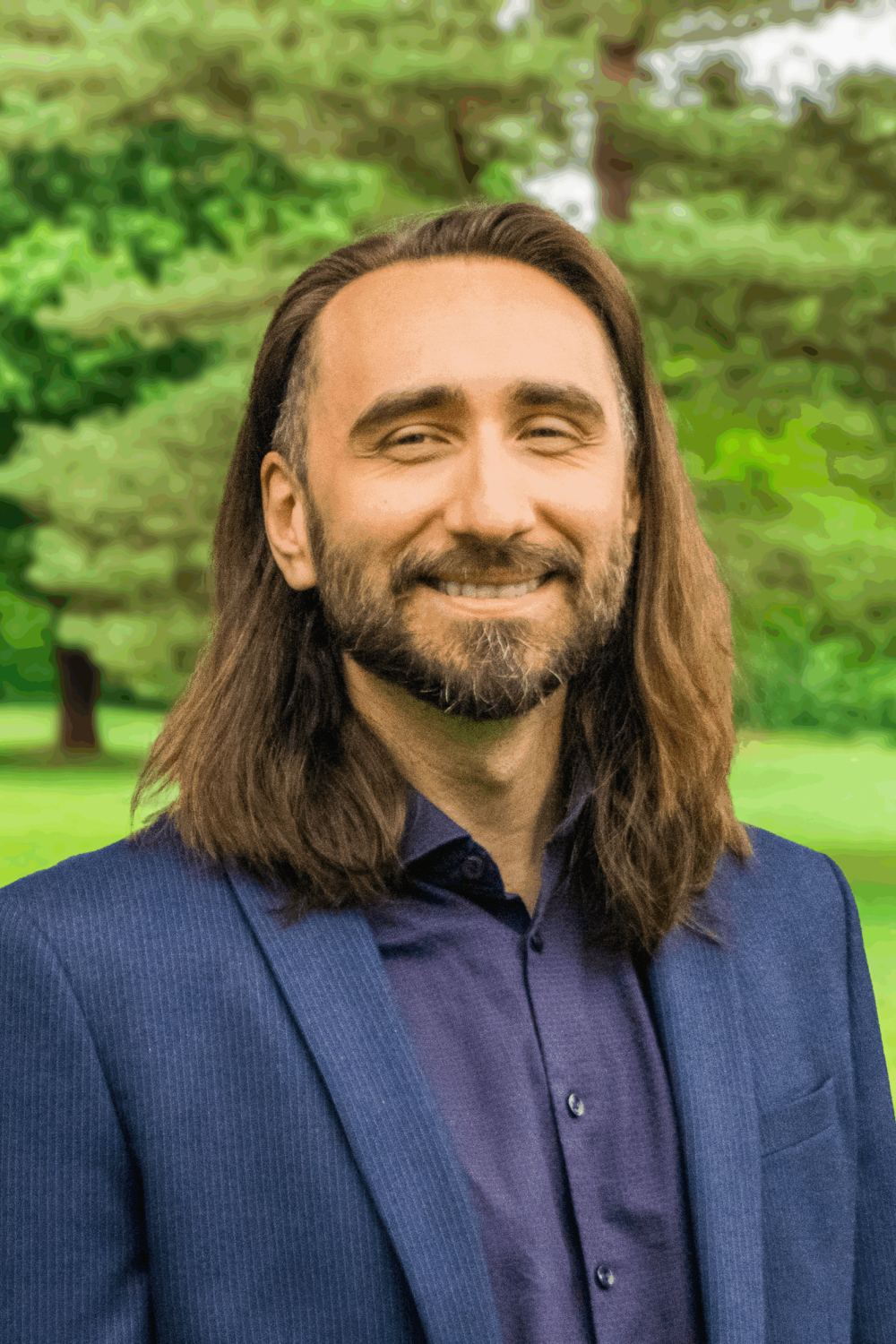 Portrait of Dr. Alexey Breuss, a man with long hair and a beard, smiling outdoors in a green park.